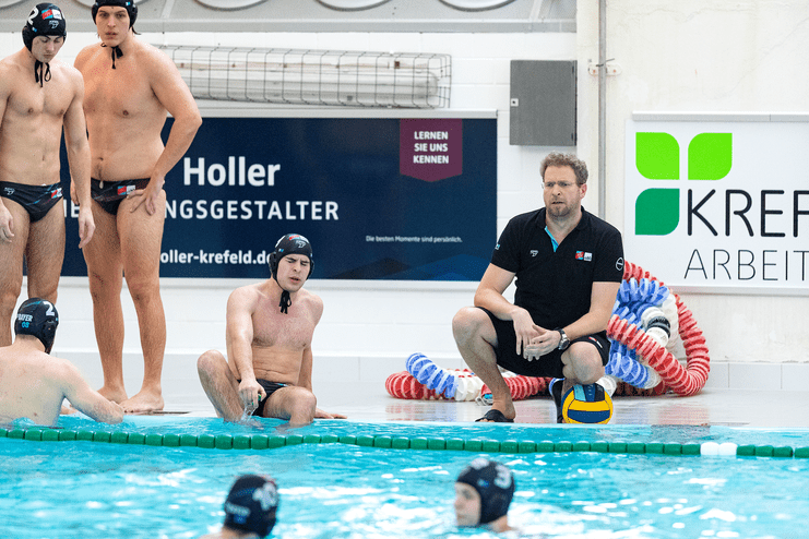 Wasserballmannschaft bei einer Besprechung am Beckenrand, Trainer mit Ball und Spieler in Schwimmkleidung.