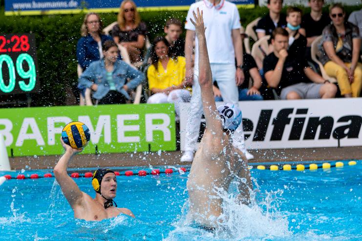 Wasserballspieler mit gelbem Ball im Sprung, während ein Gegner zum Blocken aufspringt. Zuschauer im Hintergrund.
