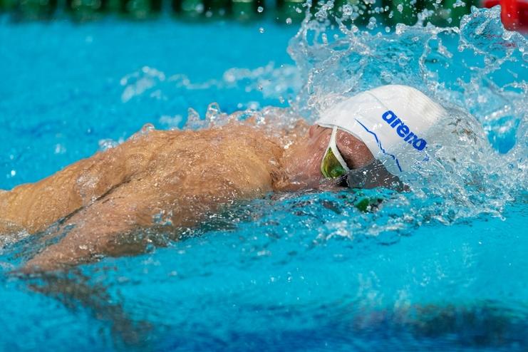 Schwimmer in weißer Badekappe und grünen Schwimmbrille taucht mit einem Arm ins Wasser, Spritzwasser um ihn herum.