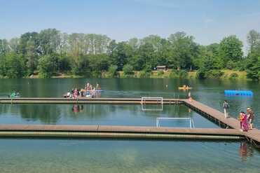 Blick auf einen Badesee mit Holzplattformen, Schwimmern, Paddelbooten und umliegendem Baumbestand.