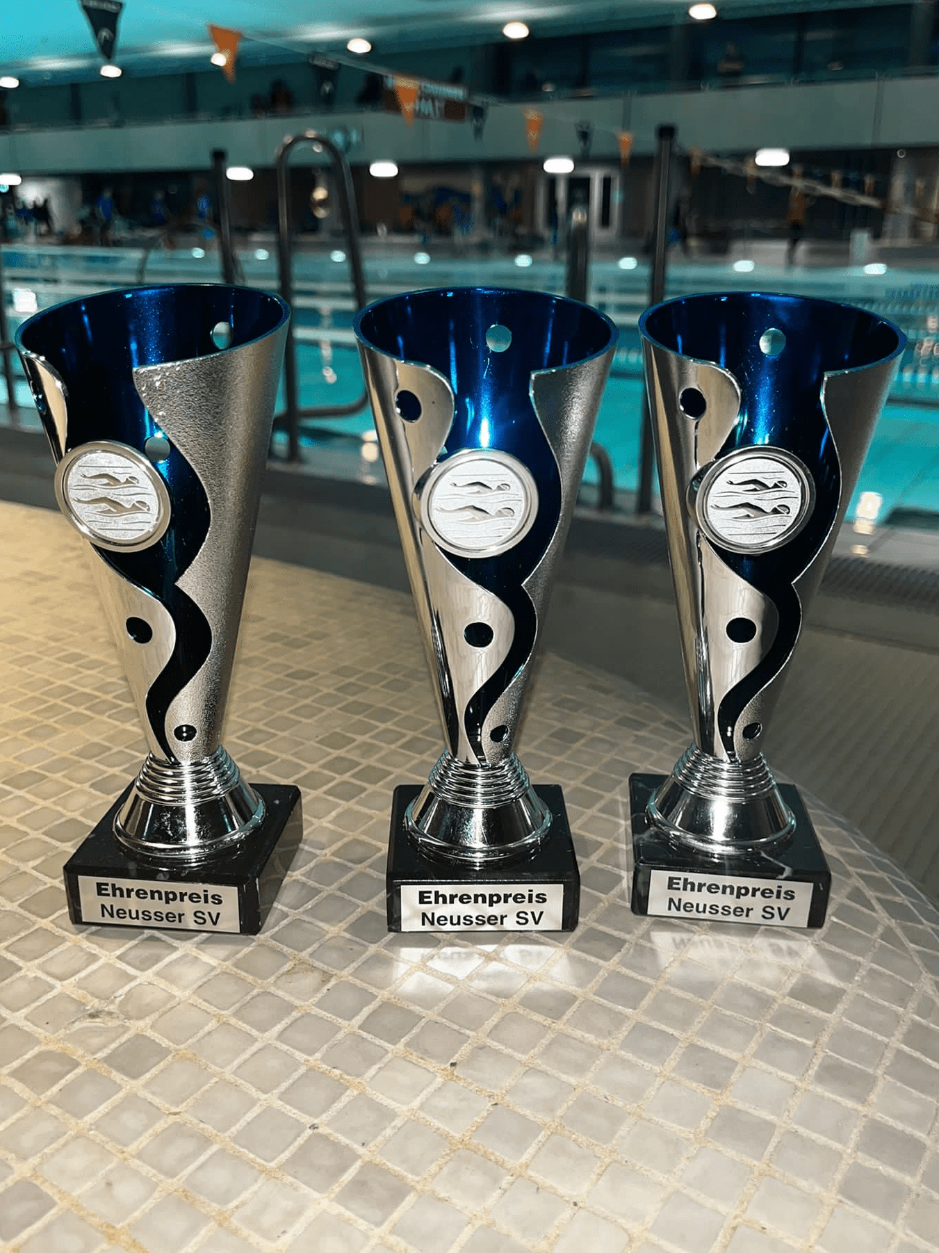 Drei silberne Pokale mit blauen Oberteilen, beschriftet mit "Ehrenpreis Neusser SV", stehen an einem Tisch neben einem Schwimmbad.