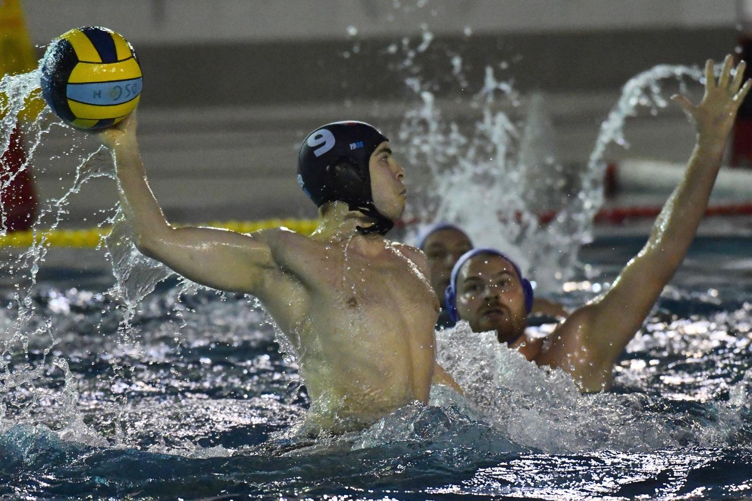 Wasserballer wirft einen Ball &uuml;ber spritzendes Wasser, w&auml;hrend ein Gegenspieler versucht, ihn zu blockieren.