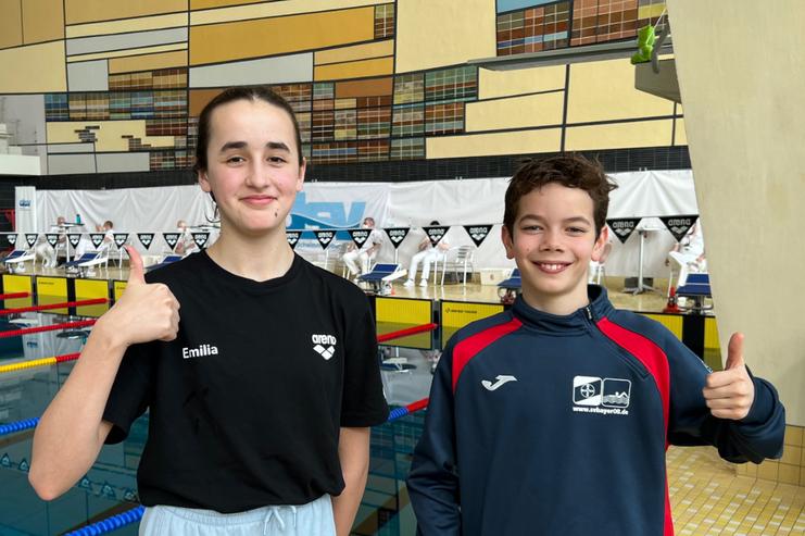 Zwei Jugendliche in Schwimmbekleidung stehen am Beckenrand und geben Daumen hoch, Hintergrund mit Schwimmbahnen.