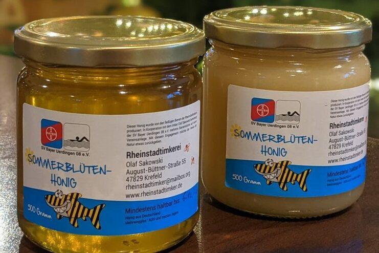 Zwei Glasbehälter mit Sommerblütenhonig, einer gold- und der andere beigefarben, mit Etiketten der Rheinstadtimkerei.