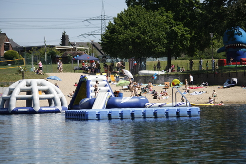 Inflatable Wasserspielplatz auf einem Gewässer, umgeben von zahlreichen Badenden und Freizeitaktivitäten am Strand.