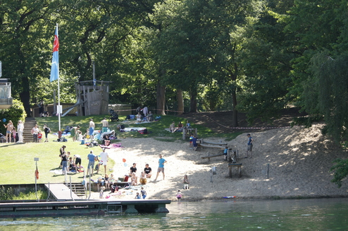 Badegäste entspannen am Sandstrand, während Kinder im Grünen und auf einem Spielplatz spielen. Bäume im Hintergrund.