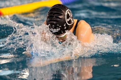 Schwimmerin in schwarzer Badekappe taucht mit gesenktem Kopf ins Wasser und erzeugt Spritzwasser.