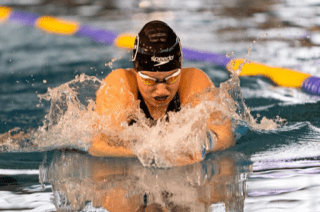 Schwimmerin in voller Aktion beim Brustschwimmen, mit Spritzwasser und bunten Bahnen im Hintergrund.