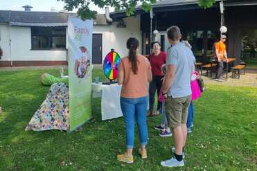 Gruppenszene im Freien mit Menschen um ein Glücksrad und einem Informationsstand unter einem Baum.