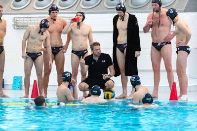 Wasserballmannschaft w&auml;hrend einer Besprechung am Beckenrand; einige Spieler im Wasser, andere stehen um den Trainer.