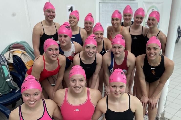Gruppe von 16 Schwimmerinnen in pinken Badekappen, posierend am Beckenrand in einem Schwimmhallenraum.