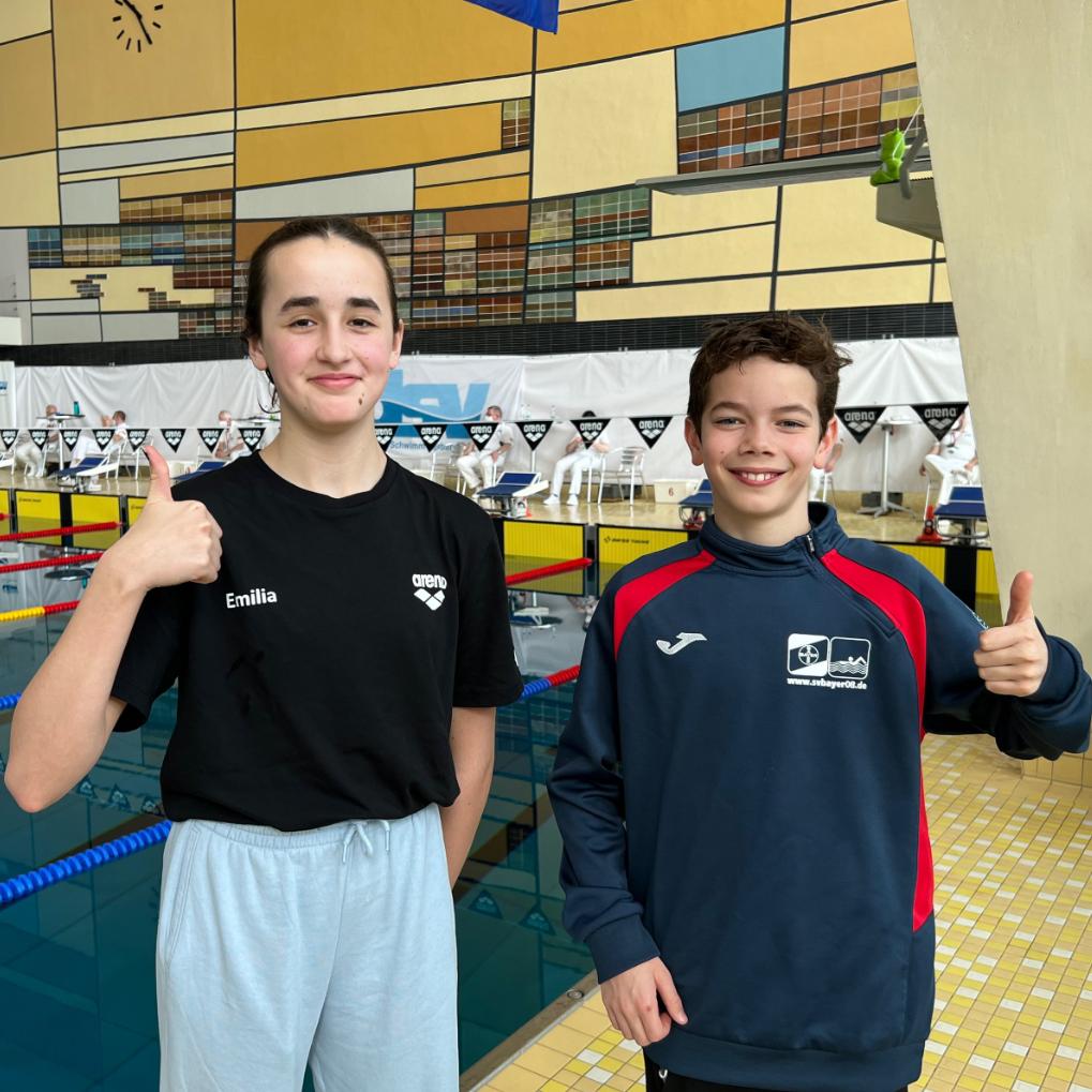 Zwei Jugendliche in Schwimmbekleidung stehen am Beckenrand und geben Daumen hoch, Hintergrund mit Schwimmbahnen.