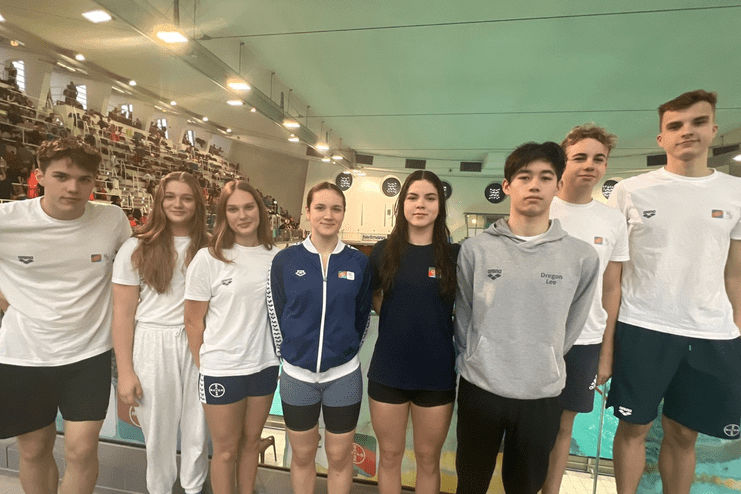 Gruppe junger Schwimmer steht nebeneinander am Beckenrand in einem Sportbad, Zuschauer im Hintergrund.