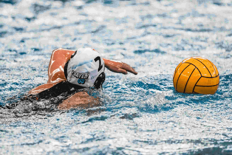 Wasserballspielerin schwimmt mit gelbem Ball im Wasser, Blick nach vorne, weiße Badehaube mit „BAYER 03“.