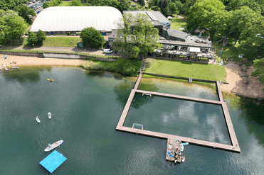 Luftaufnahme eines Gewässers mit Steg, Freizeitaktivitäten und Gebäuden in einem grünen Umfeld.