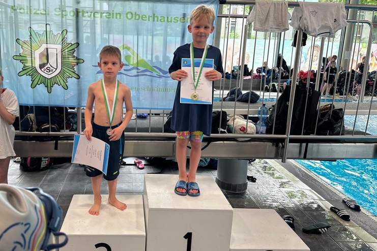 Zwei Kinder stehen auf dem Treppchen bei einem Schwimmwettkampf, der eine Medaille und eine Urkunde hält.