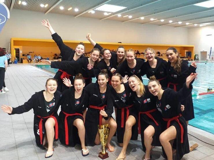 Gruppe von Jugendlichen in schwarzen Bademänteln mit Pokal, lächelnd am Beckenrand eines Schwimmbads.