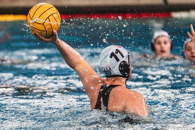 Wasserballspielerin mit gelbem Ball im Wurf, im Hintergrund sind Mitspielerinnen im Wasser zu sehen.