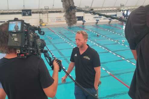 Mann steht am Beckenrand eines Schwimmbads, w&auml;hrend eine Kamera und ein Mikrofon auf ihn gerichtet sind.