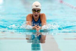 Schwimmerin in blauer Badebekleidung mit Badekappe und Schutzbrille vollzieht einen Brustschwimmzug im Wasser.