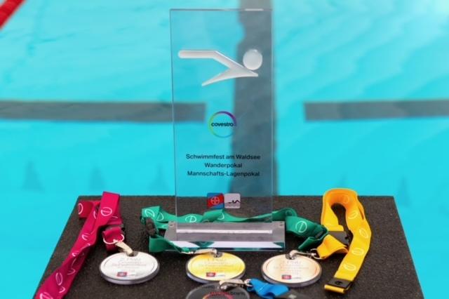 Pokale und Medaillen für ein Schwimmfest, platziert auf einem Tisch vor einem Schwimmbecken.