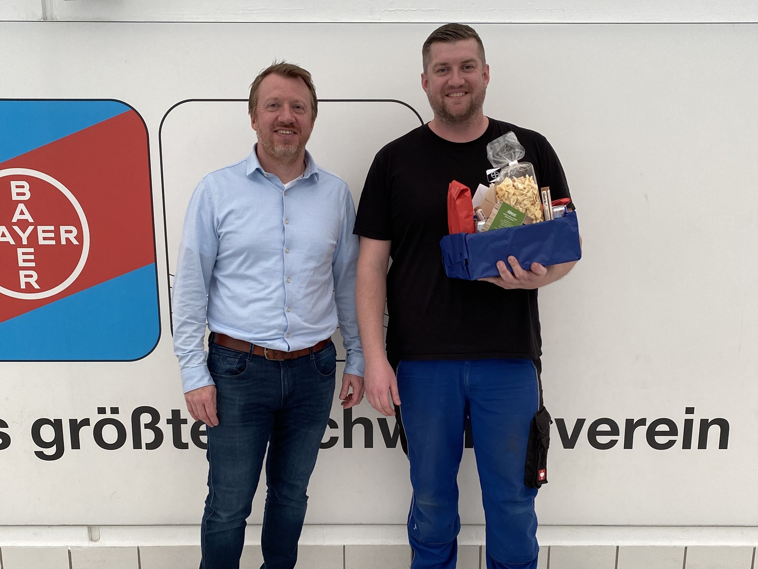 Zwei Männer stehen vor einer Wand mit dem Bayer-Logo, einer hält ein Geschenk, der andere lächelt.