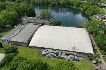 Luftaufnahme eines Sportgebäudes mit einer weißen Kuppel, umgeben von Bäumen und einem nahegelegenen Gewässer.