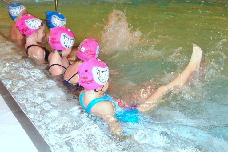 Sechs Kinder in bunten Badekappen sitzen im flachen Wasser eines Hallenschwimmbads und spritzen fröhlich.