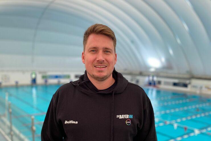 Mann in schwarzem Hoodie steht vor einem Schwimmbecken in einer Hallenbad-Anlage mit Menschen im Wasser.