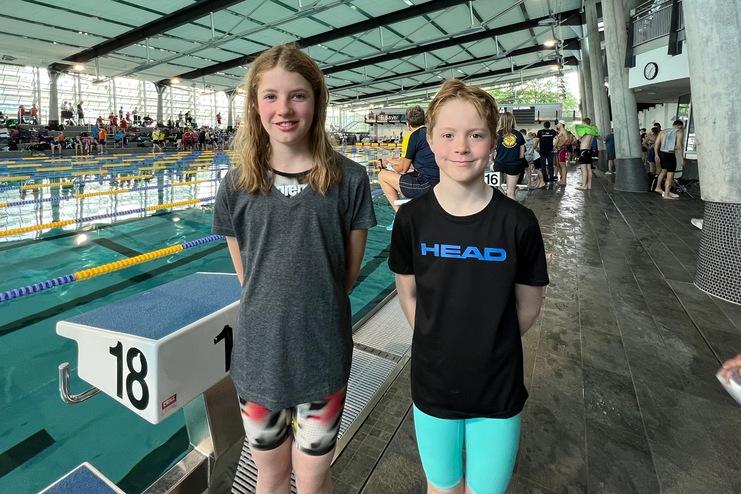 Zwei Kinder lächeln vor einer Schwimmhalle, im Hintergrund sind Schwimmbecken und Zuschauer zu sehen.