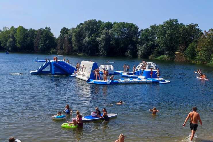 Badeszenario an einem See mit Menschen, Spielgeräten und Sand am Ufer; Aktivitäten im Wasser und am Strand sichtbar.