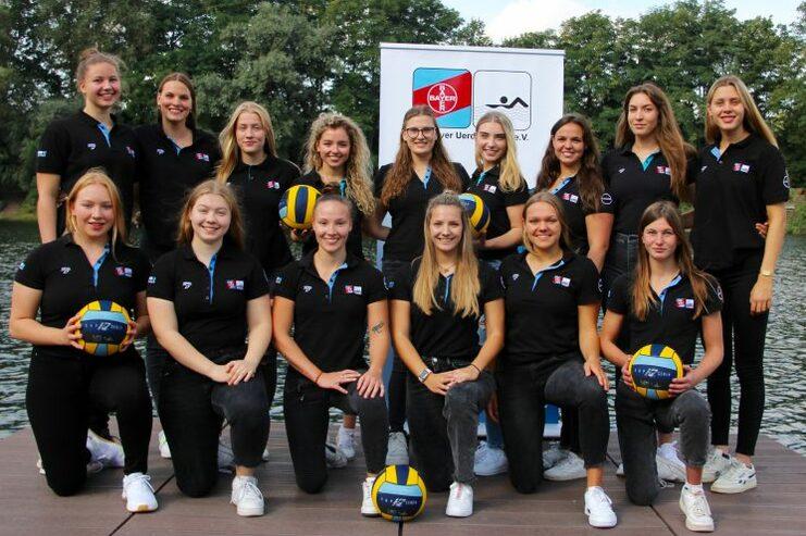 Gruppenbild von 15 weiblichen Volleyballspielerinnen in schwarzen Poloshirts, einige halten Bälle, hinter ihnen ein Logo.