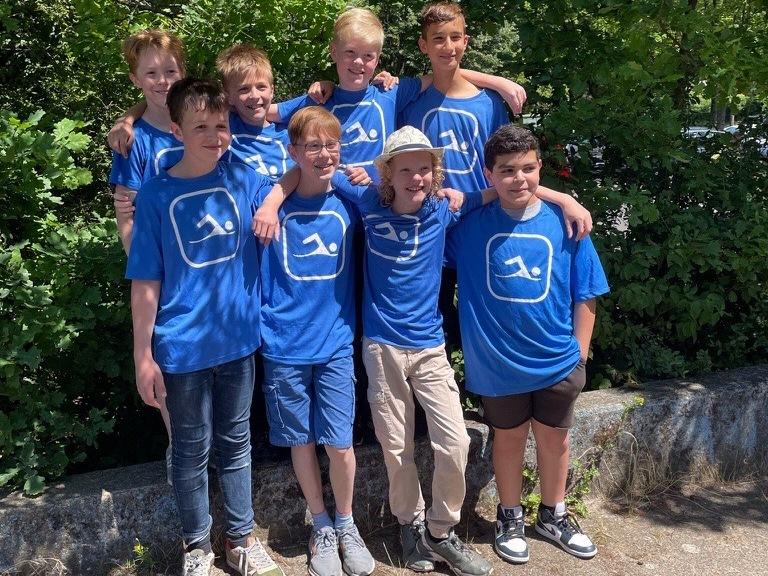 Gruppe von acht Jungen in blauen T-Shirts mit Logo, stehen l&auml;chelnd vor einer gr&uuml;nen Alternative mit Holzbank.