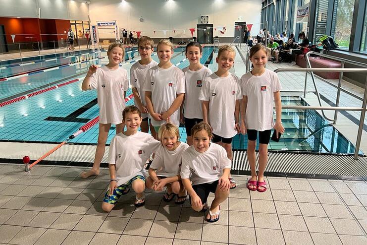 Gruppennfoto von zehn Kindern in weißen T-Shirts am Beckenrand eines Schwimmbads, lächeln und posieren.