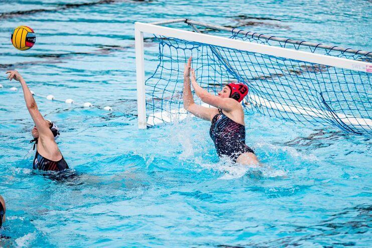 Wasserballspiel: Spielerin springt, um einen Ball vor dem Tor zu blocken, während sie im Wasser steht.