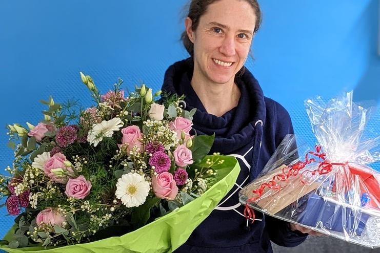 Frau mit einem großen Blumenstrauß und einem Geschenk in der Hand steht vor einer blauen Wand, auf einem grauen Sofa.