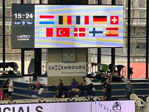 Digitale Anzeigetafel mit Flaggen verschiedener Länder und Zeitangabe in einem Schwimmbad. Zuschauer und Sportler auf Tribüne.