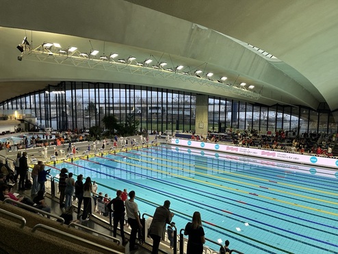 Blick auf ein Hallenbad mit Schwimmern und Zuschauern, leuchtende Werbung an der Wand, hohe Fenster und Pflanzen im Hintergrund.