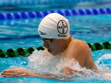 Junger Schwimmer mit Badehaube und Schwimmbrille, der kräftig in einem Becken mit Spritzwasser schwimmt.