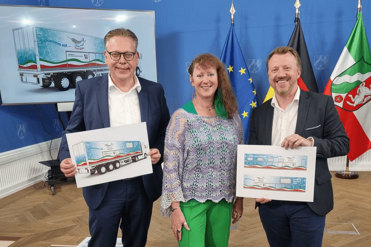 Drei Personen halten Plakate mit einem bunten Lkw-Design in einem Raum mit Flaggen und Präsentationsbild.