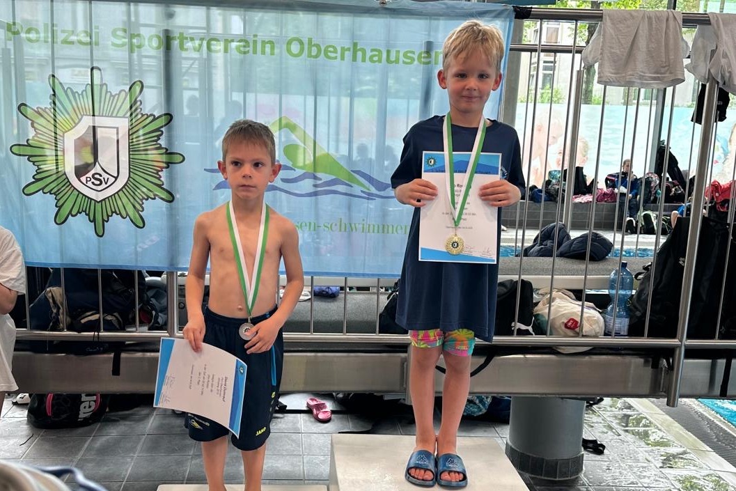 Zwei Kinder stehen auf dem Treppchen bei einem Schwimmwettkampf, der eine Medaille und eine Urkunde hält.