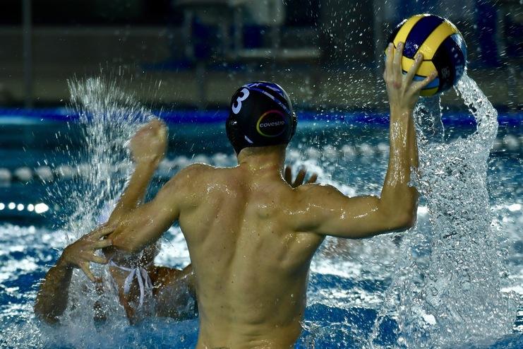 Wasserballspieler wirft einen Ball, während er von einem Gegner im Wasser gestört wird. Spritzwasser ist sichtbar.