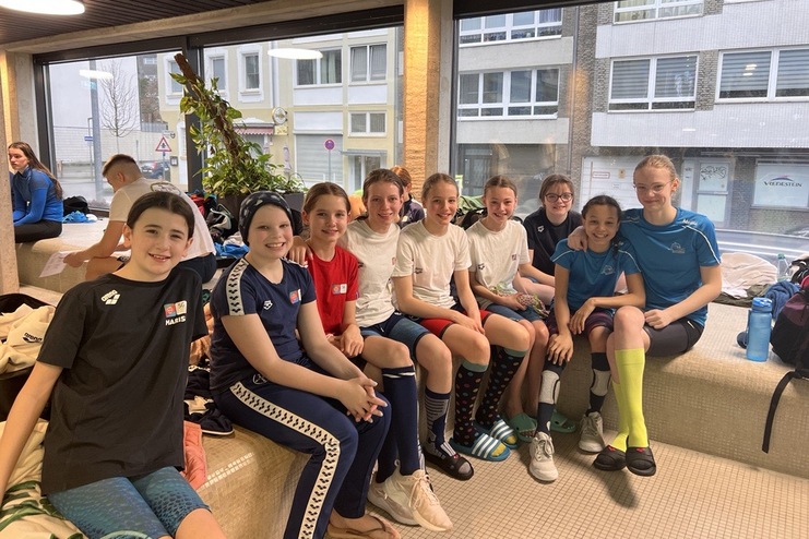 Gruppenfoto von acht Mädchen in Sportkleidung, die auf einem Sitzbereich in einem hellen Raum posieren.