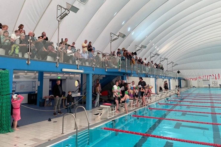 Innenaufnahme eines Schwimmbeckens mit Zuschauern auf einer Tribüne und Schwimmern am Beckenrand.