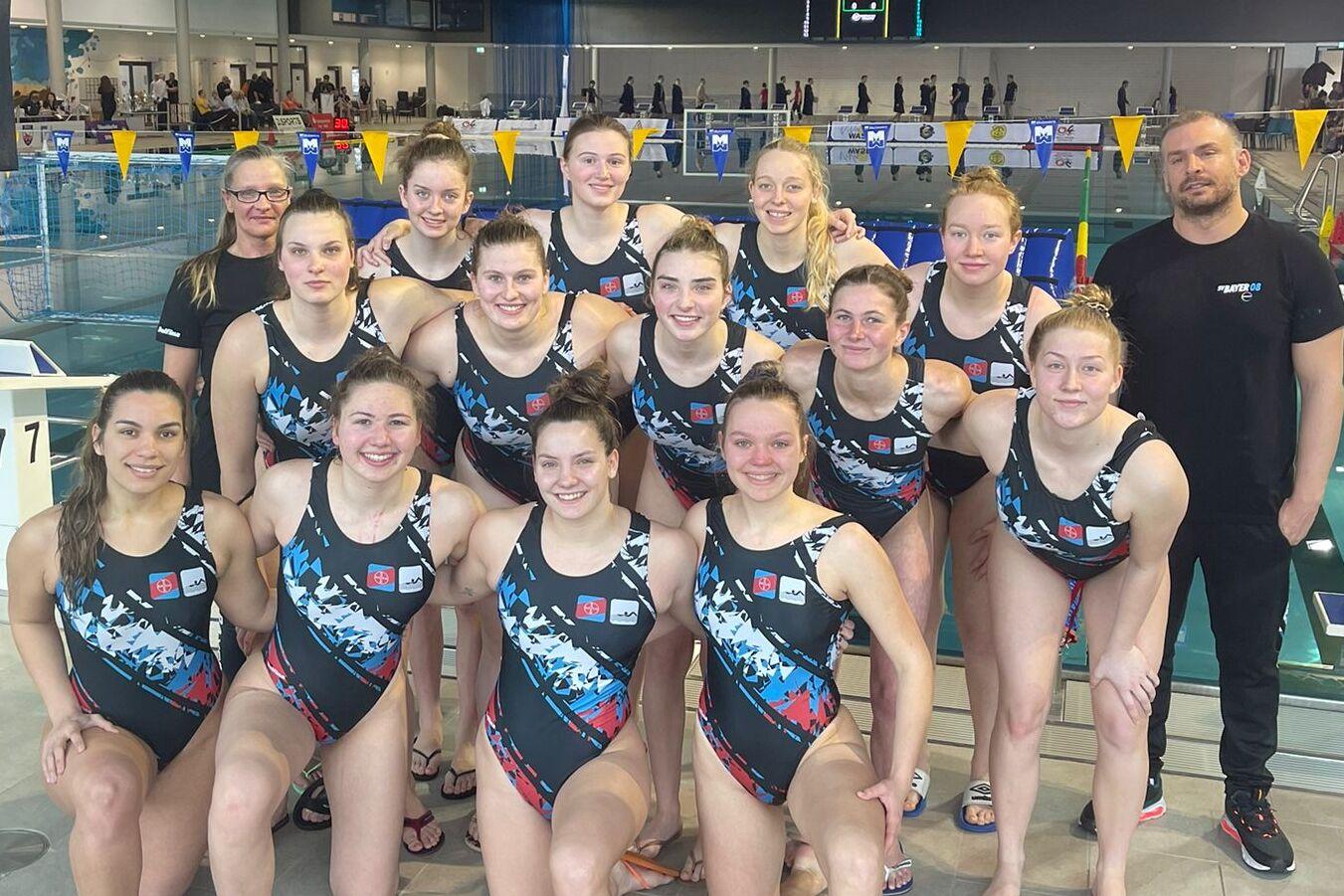 Gruppenfoto von 14 Schwimmerinnen in Wettkampfanz&uuml;gen, posierend am Schwimmbecken mit Trainer im Hintergrund.