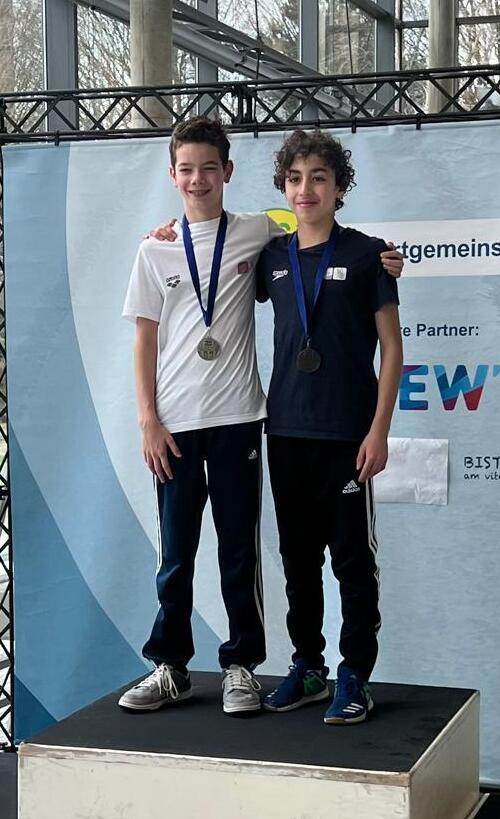 Zwei junge Sportler stehen auf dem Siegerpodest, tragen Medaillen und l&auml;cheln in die Kamera.