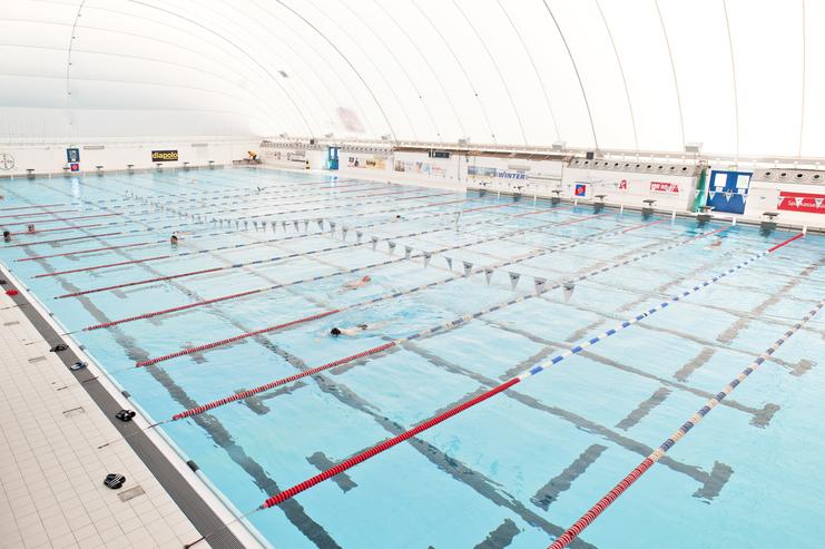 Helle Schwimmhalle mit mehreren Schwimmbahnen, in denen Schwimmer aktiv sind, unter einer aufblasbaren Decke.