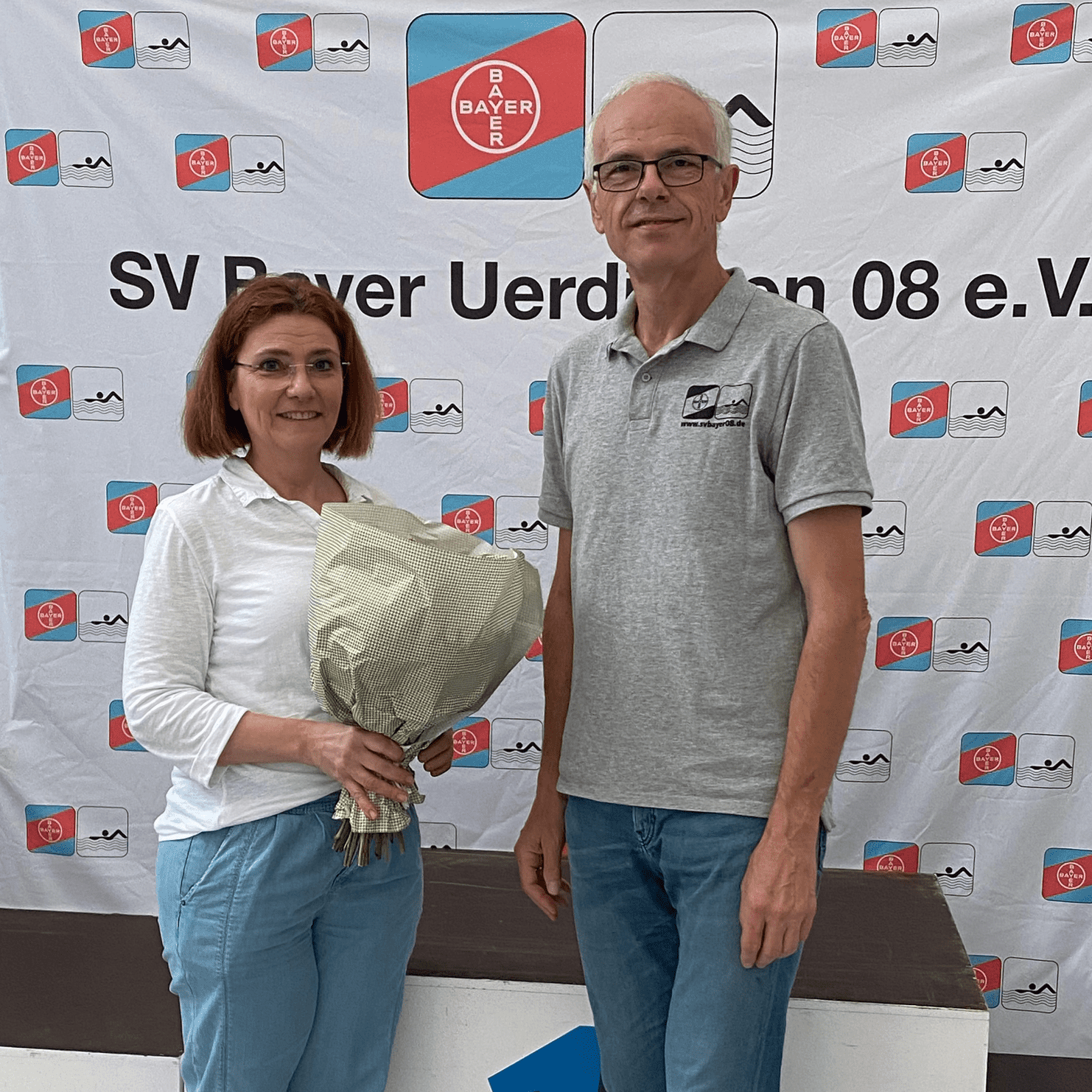 Zwei Personen stehen vor einer Sponsorenwand und halten einen Blumenstrau&szlig;. Im Hintergrund ist ein Siegertreppchen sichtbar.