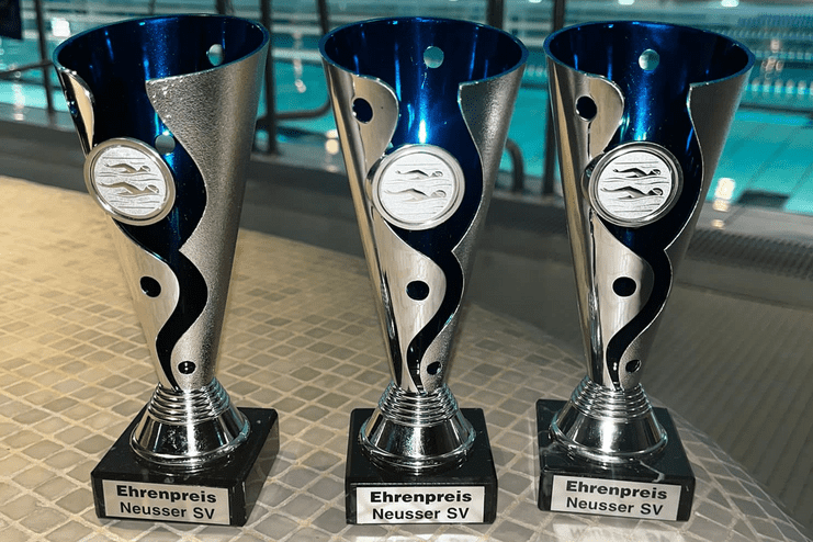 Drei silberne Pokale mit blauen Oberteilen, beschriftet mit "Ehrenpreis Neusser SV", stehen an einem Tisch neben einem Schwimmbad.