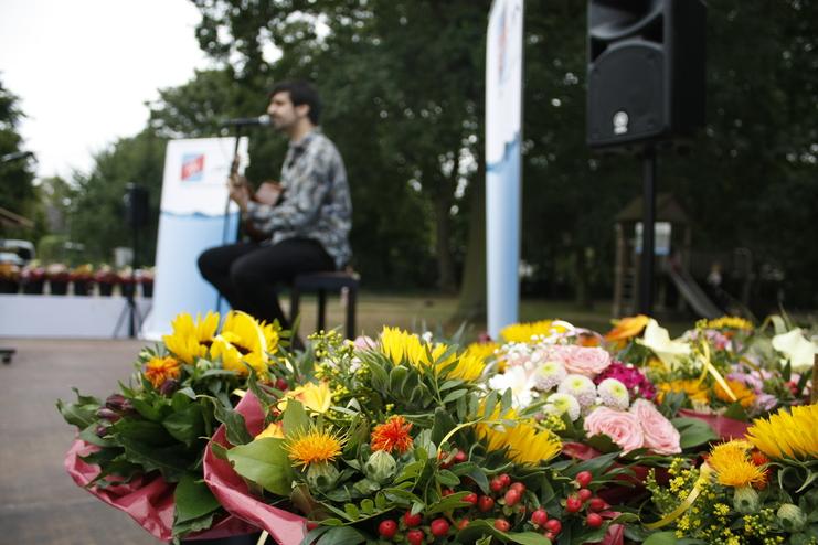 Musiker mit Gitarre auf einer Bühne, umringt von bunten Blumensträußen und einem Lautsprecher im Hintergrund.